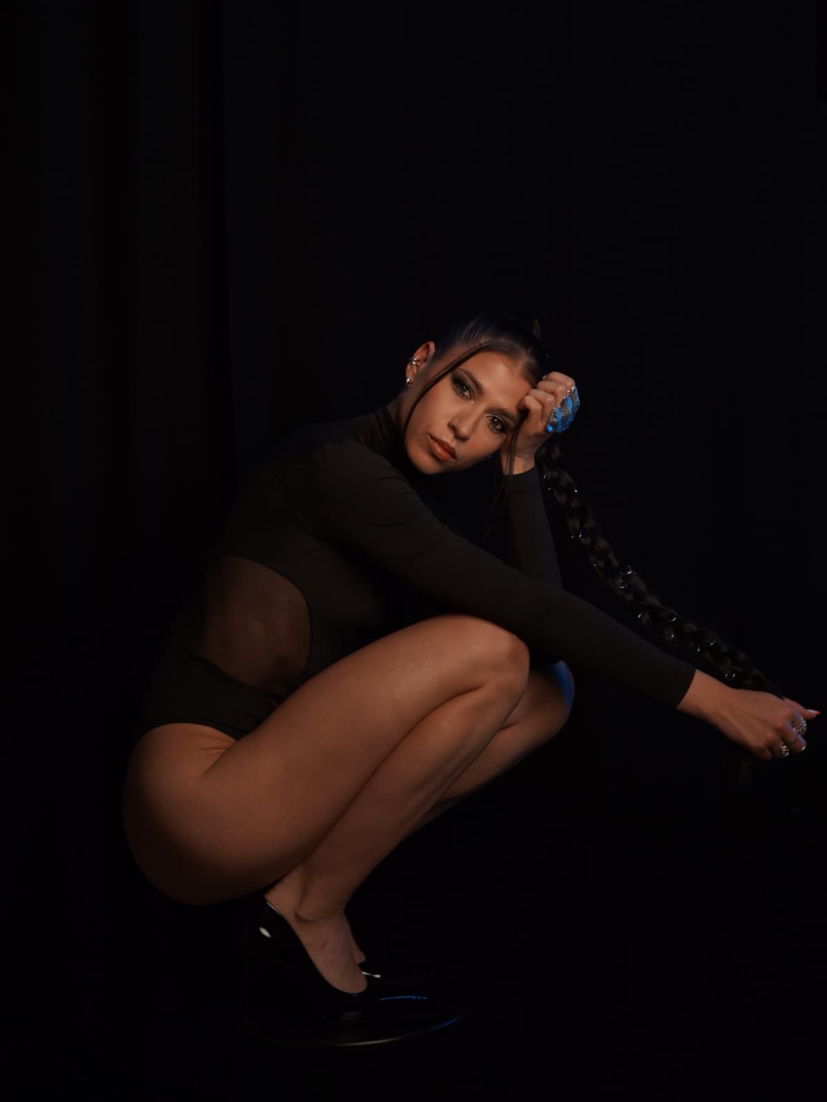 Yana squatting in a black bodysuit, wearing a heel